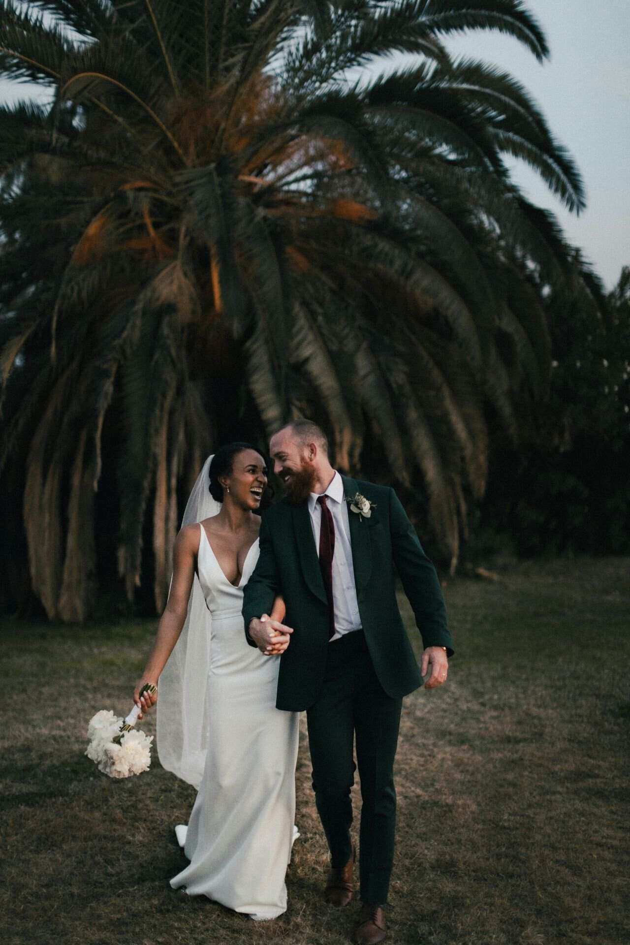 Elegant wedding couple walking outdoors with palm trees in the background.